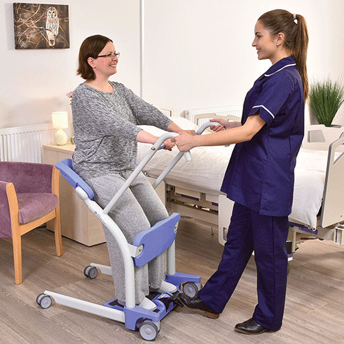 Kvinde sidder i Sara Stedy. Anden kvinde skubber kvinde i Sara Stedy hen til stol, hvor hun skal hjælpes ned i ved hjælp af Sara Stedy.